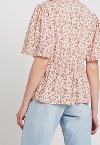 Kukkakuvioinen paita vaaleanpunaisena, jossa on lyhyet puffihihat ja kerätty vyötärö. Valmistettu pehmeästä kankaasta, jossa on herkkä pieniä kukkia esittävä kuvio.