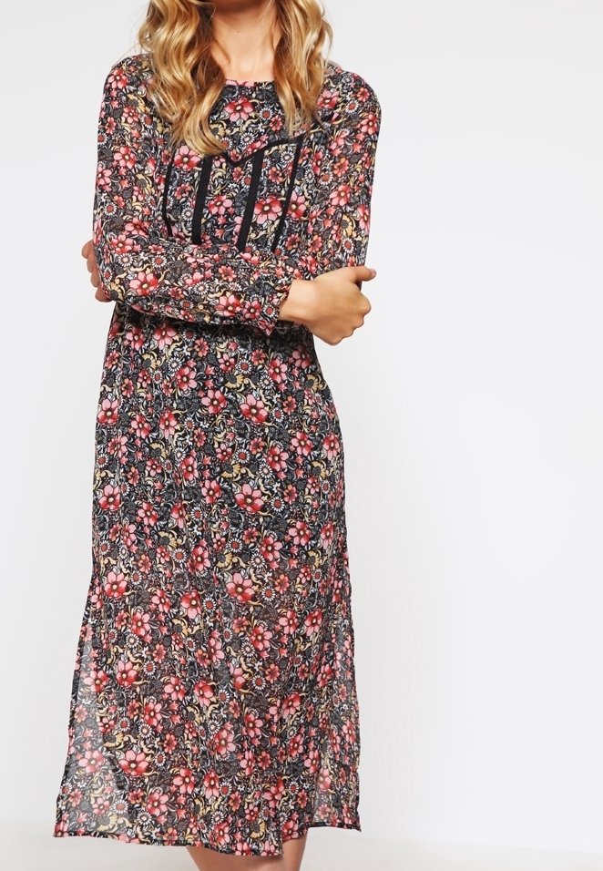 Femme aux bras croisés portant une robe fleurie à manches longues, longueur cheville, en rouge, rose et noir, avec une bordure verticale noire sur la poitrine.