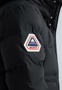 Čierna nafukovacia bunda s hexagonálnou bielou nášivkou s červenými a modrými detailmi so slovom "JACKiT" na ľavom rukáve. Textúrovaná tkanina.