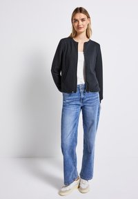 Kvinna iklädd en svart vadderad jacka med dragkedja över en vit topp, blå raka jeans och vita sneakers stående mot en enkel vit bakgrund.