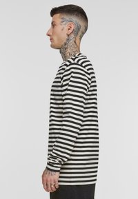 Chemise à manches longues rayée noir et blanc avec un col rond et une coupe décontractée. Des rayures horizontales s'étendent des épaules à l'ourlet.