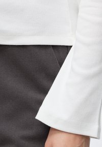Gros plan d'une personne portant un haut blanc côtelé à manches longues et un pantalon gris foncé avec une poche visible et un poignet de manche.