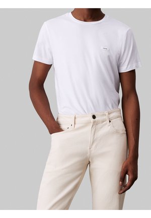 Man in een witte Calvin Klein T-shirt, ingestopt in hoge, off-white jeans, staande met één hand in zijn broekzak.