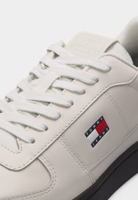 Λευκό αθλητικό παπούτσι με λευκά κορδόνια, μαύρη σόλα και κεντημένη ετικέτα με το λογότυπο Tommy Jeans στο πλάι.