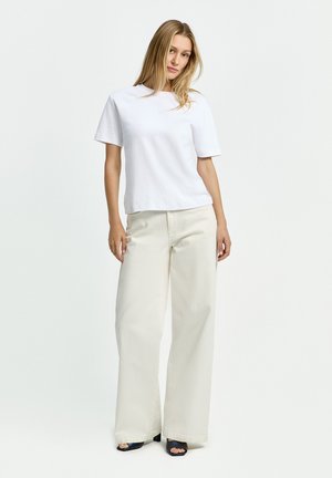 Frau steht und trägt ein schlichtes weißes T-Shirt mit kurzen Ärmeln, cremefarbene Weitbein-Hose und schwarze offene Absatzschuhe vor weißem Hintergrund.