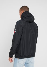 Svart hettejakke med elastisk liv, med et logo-patch på venstre erme, og et glatt, vannavstøtende stoff.