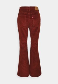 Pantalon évasé en velours côtelé couleur rouille, présentant une taille haute, deux poches arrière, et une étiquette en cuir sur la ceinture. Tissu texturé.