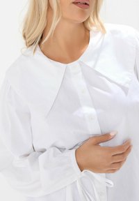 Biała bluzka z dużym, spiczastym kołnierzem, zapinana na guziki z przodu oraz obszerne rękawy z elastycznymi mankietami. Gładka tekstura, minimalistyczny design.