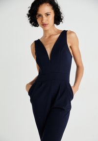 Μπλε ναυτικό ολόσωμο φόρεμα με βαθύ V, αμάνικη σχεδίαση, τσέπες στα πλαϊνά και εφαρμοστό μπούστο που καταλήγει σε χαλαρά μπατζάκια. Λείο ύφασμα.