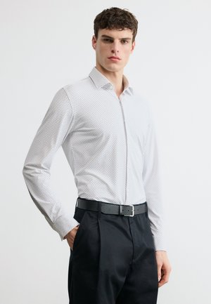 Young man wearing a white patterned long-sleeve shirt tucked into black pants with a black belt, standing against a plain background.