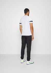 Witte poloshirt met zwarte streepaccenten, gecombineerd met zwarte trainingsbroek met drukknoopjes langs de zijkanten en witte sportschoenen.