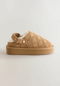 Beige Slipper mit einer strukturierten Wildleder-Oberfläche, ausgestattet mit einem weichen Kunstpelzfutter, einer gepolsterten Plattform und einem verstellbaren Knöchelriemen mit einem goldfarbenen Knopf.