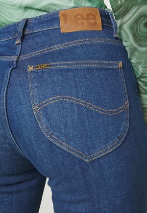 Nahaufnahme der blauen Lee-Jeans-Hintertasche mit orangefarbenen Nähten und Leder-Brandpatch am Bund, getragen mit einem grünen Oberteil aus strukturiertem Stoff.