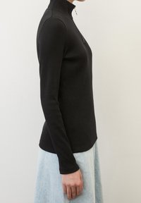 Maglietta nera a coste con maniche lunghe, colletto alto e chiusura con zip, abbinata a una gonna di jeans chiara, esposta di profilo.