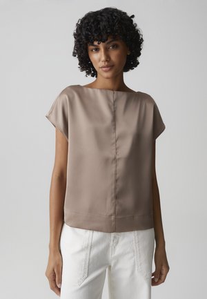 Frau mit lockigem Haar, die eine kurzärmelige Bluse aus Satin in Taupe und weiße Hosen trägt, vor einem schlichten hellgrauen Hintergrund steht.