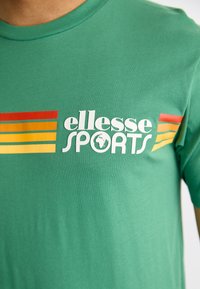 Grøn bomulds T-shirt med et stort hvidt "ellesse SPORTS" logo, accentueret af horisontale striber i rød, gul og orange.