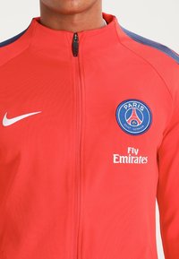 Punainen vetoketjullinen takki, joka on valmistettu sileästä kankaasta ja jossa on siniset yksityiskohdat sekä Paris Saint-Germain -logo rinnassa. Takissa on myös 'Fly Emirates' -teksti.