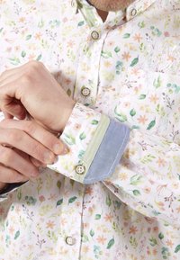 Chemise à boutonnières florale avec une base blanche ornée de fleurs et de feuilles pastel. Les poignets présentent des accents vert clair et bleu avec des coutures contrastées.