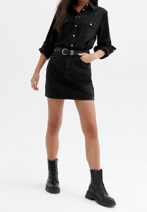 Femme portant une chemise noire boutonnée, une mini-jupe noire avec ceinture, et des bottes de combat noires à lacets, debout avec une main dans la poche.