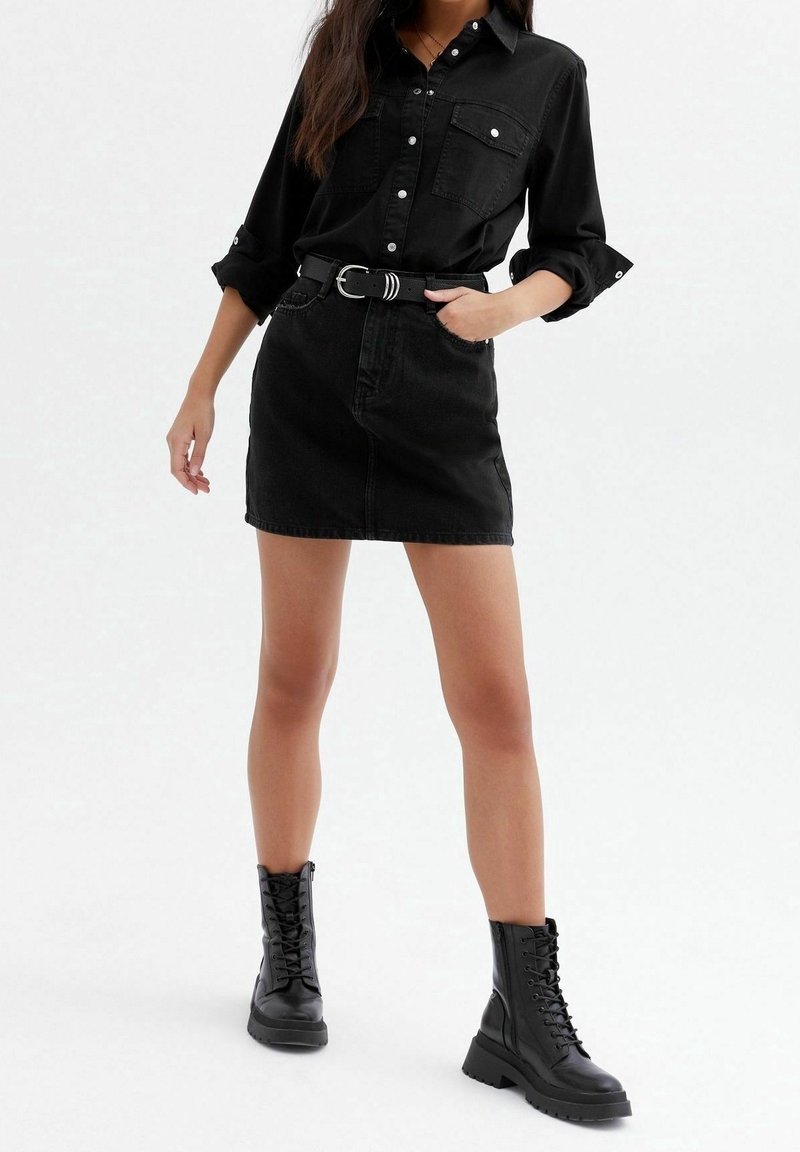 Femme portant une chemise noire boutonnée, une mini-jupe noire avec ceinture, et des bottes de combat noires à lacets, debout avec une main dans la poche.