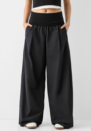 Pantalones negros de pierna ancha con un tejido suave y ligero, que cuentan con una cinturilla alta elástica y bolsillos laterales, combinados con zapatos blancos.