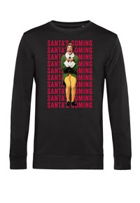 Felpa nera a maniche lunghe con una grande grafica di un personaggio in un abito verde con accenti bianchi, circondato da testo ripetuto: "SANTA STA ARRIVANDO."