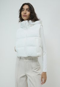 Giovane donna con lunghi capelli scuri che indossa un gilet bianco imbottito sopra una maglia a maniche lunghe bianca e pantaloni beige a vita alta su uno sfondo semplice.