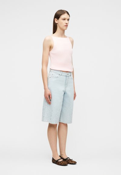 Γυναίκα που στέκεται, φοράει απαλό ροζ αμάνικο τοπ, ανοιχτό μπλε τζιν σορτς μέχρι το γόνατο με πιτσιλιές μπογιάς και μπαλαρίνες με λεοπάρ σχέδιο.