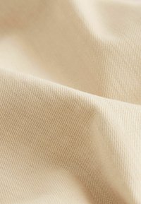 Helles beige Gewebe mit glatter Textur, das ein diagonales Rippenmuster aufweist. Das Material ist flexibel und zeigt in den Falten einen dezenten Glanz.