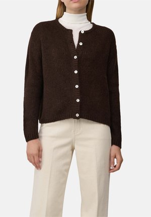 Femme portant un cardigan marron foncé boutonné sur un haut blanc à col montant et un pantalon couleur crème sur un fond uni clair.