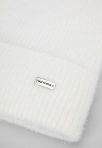 Gorro blanco de punto con textura acanalada y una etiqueta metálica lisa con la palabra "WITTCHEN" adherida al borde.