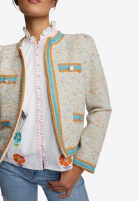 Veste texturée multicolore avec des accents orange et bleu. Comprend des boutons métalliques et deux poches avant, associée à une chemise brodée.