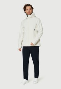 Herrernas vita vattentäta jacka med huva, med svarta dragkedjefickor och svarta detaljer, kombinerad med marinblå byxor och vita sneakers.