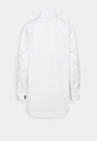 Chemise blanche à manches longues boutonnée, vue de dos, avec col et un petit détail noir en forme de lettre sur le poignet gauche.