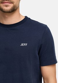 Marineblå bomulls t-skjorte med rund hals og korte ermer. Viser ordet "JEFF" i hvit brodering på venstre brystområde.