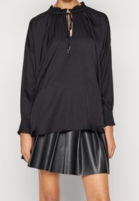 Czarna bluzka z długim rękawem, z marszczonym dekoltem i sznurkiem, zestawiona z plisowaną czarną skórzaną spódnicą. Gładka faktura tkaniny, luźny krój.