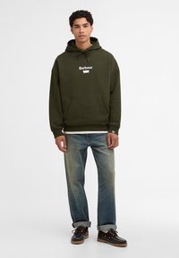 Ελιά πρασίνου φόρμα με μπροστινή τσέπη και λευκό λογότυπο "Barbour". Συνδυασμένο με ανοιχτόχρωμα μπλε τζιν και σκούρα καφέ παπούτσια με ανθεκτική υφή.