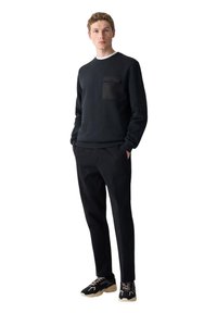 Zwart katoenen sweatshirt met een contrasterende zak, gecombineerd met zwarte broeken en zwarte sneakers met beige accenten. Neutrale achtergrond.