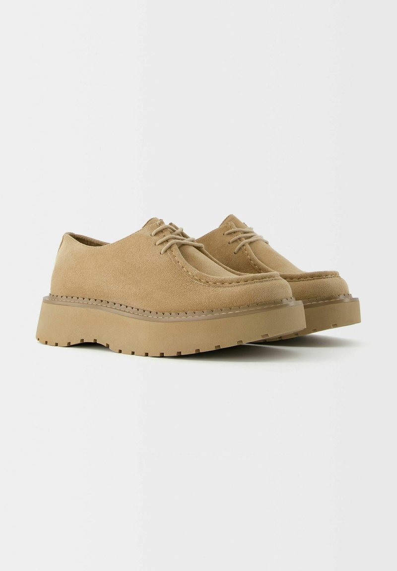Zapatos de ante beige con punta redondeada y suela de plataforma resistente. El diseño simple de cordones presenta costuras en tono y detalles texturizados en la suela.