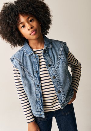 Denimvest med ærmeløs design, lys blå vask og knaplukning foran. Underneath en stribet langærmet skjorte i beige og marineblå.