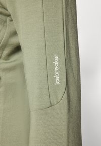 Camisa verde de mangas longas com uma textura suave e um design ajustado. Apresenta costuras cosidas e o logotipo "icebreaker" em bordado branco na manga.