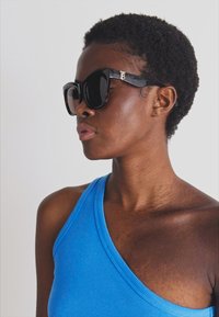 Lunettes de soleil noires surdimensionnées avec une finition brillante et un détail de logo doré sur la branche. Portées avec un haut sans manches bleu.
