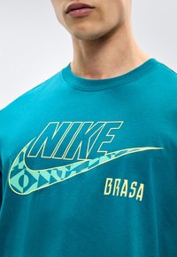 Κοντινό πλάνο ενός ατόμου που φοράει μπλουζάκι Nike βεραμάν με λογότυπο με σχέδιο και τη λέξη "BRASA" τυπωμένη από κάτω.