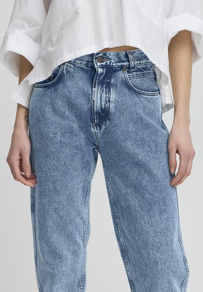 Jeans de mezclilla de lavado claro con un corte holgado, diseño de cinco bolsillos y un ligero desgastado. Combinados con una camisa blanca corta con botones.