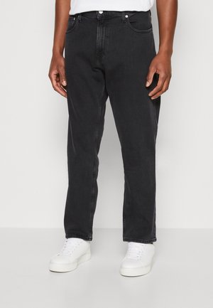 Mann trägt gerade geschnittene schwarze Jeans und weiße Turnschuhe, steht vor einem einfarbigen hellgrauen Hintergrund.