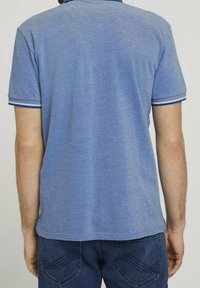 Ljusblå polo med textilstruktur, med krage och korta ärmar med blå accenter vid ärmsluten, visad bakifrån.