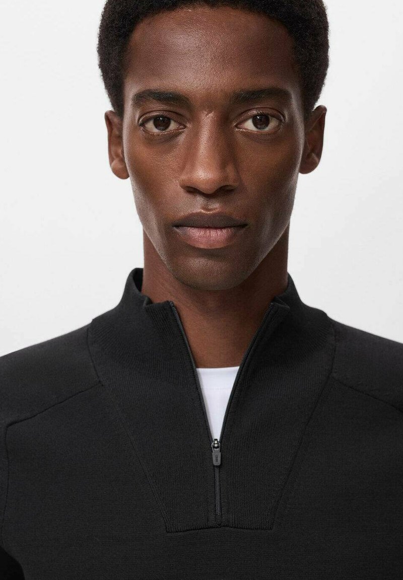 Zwarte zip-up sweatshirt met hoge kraag, met een gladde textuur en subtiele naaddetails. Draagt over een wit T-shirt.
