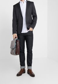 Mörkgrå blazer över en vit skjorta med geometriskt mönster, mörk denimjeans, bruna läderboots och en tvåtonad budväska.