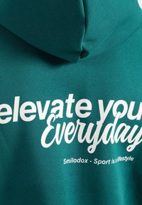 Turkusowy hoodie o miękkiej fakturze z białym napisem: "elevate your Everyday" oraz "Smilodox - Sport to styl życia" na plecach.