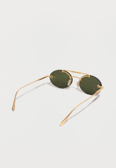 Στρογγυλά γυαλιά ηλίου με σκούρους πράσινους φακούς, λεπτά χρυσά μεταλλικά πλαίσια και μαξιλαράκια μύτης που ακουμπούν σε λευκή επιφάνεια.